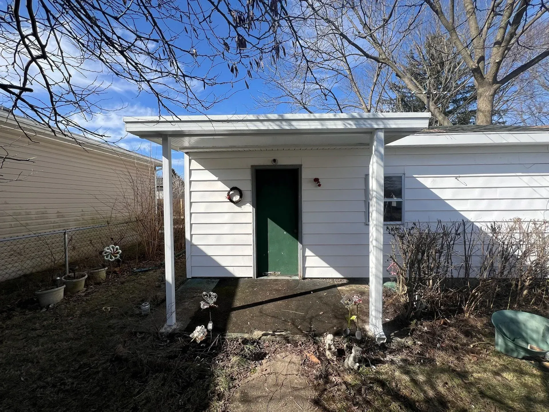 Small white building with green door under a white canopy. Blue sky and bare trees in the background.