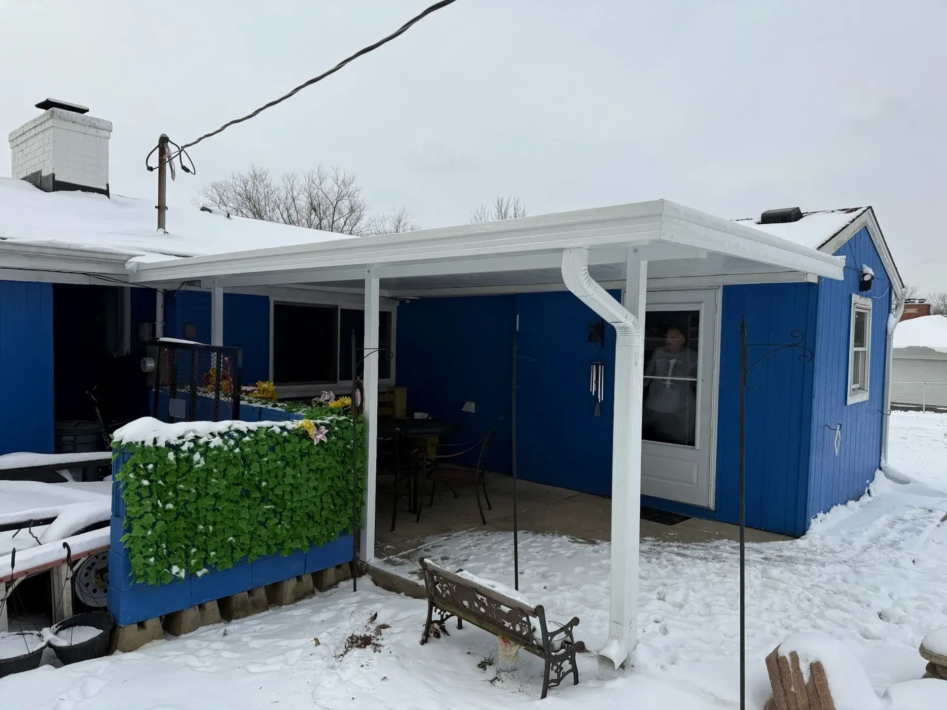 Blue house with a white-roofed patio, snow on the ground. A person is visible in the doorway.