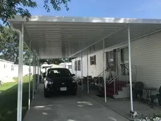 Black car parked under a white carport next to a single-story mobile home on a sunny day.