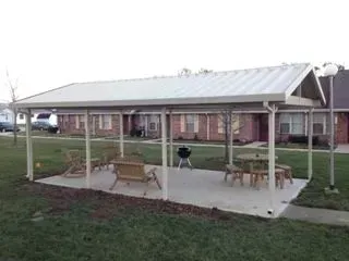 Covered outdoor patio with seating and grill. White metal frame and roof, on a concrete slab in front of brick buildings.