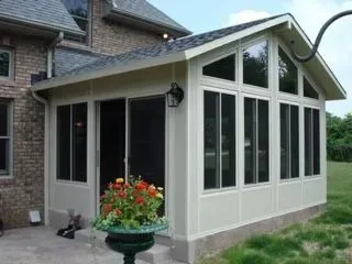 Beige-colored sunroom addition attached to a brick house, featuring screened windows and a decorative flower pot.