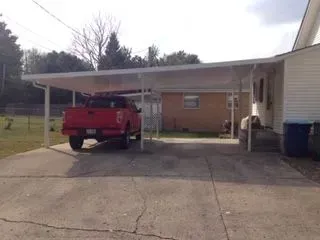 Red truck parked under a white carport attached to a beige house, on a concrete driveway.
