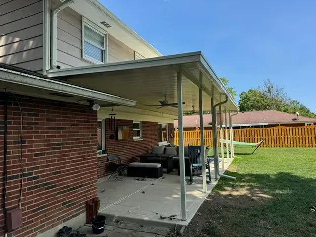 Covered patio attached to a brick house with outdoor furniture under a light-colored roof. Green lawn in background.