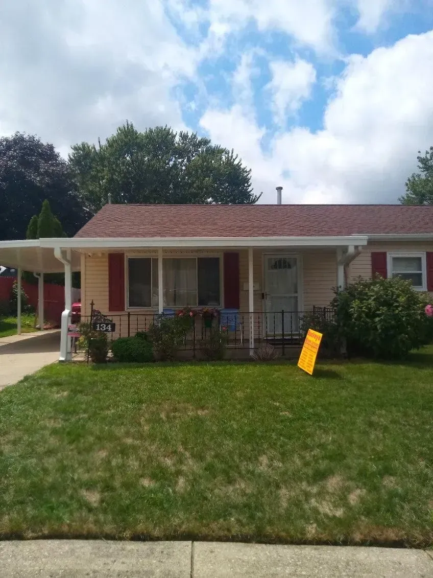 Small beige house with red shutters, carport, and yellow sign on the lawn, under a partly cloudy sky.