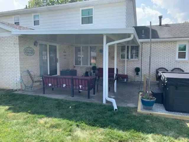 Backyard patio with white awning, seating, and hot tub near brick house with green grass.