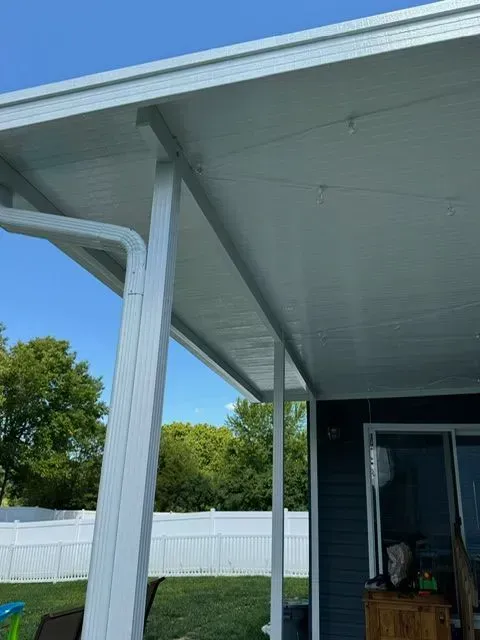 White patio roof with supporting columns, blue sky, and a glimpse of a yard and house.