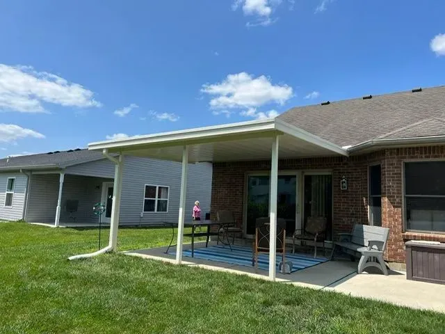 White-framed patio cover attached to a brick house, over a patio with a table and chairs, on a sunny day.