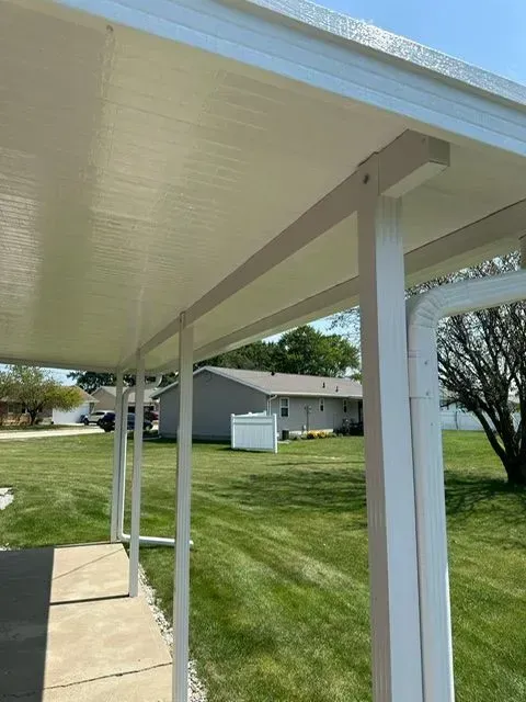 White carport with a corrugated ceiling, supported by white posts, next to a house with green grass.