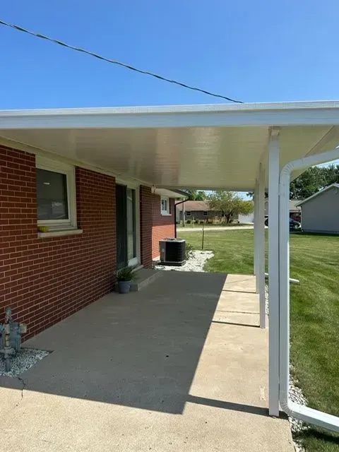 Covered porch attached to a red brick house with a concrete floor, surrounded by green grass and a clear blue sky.