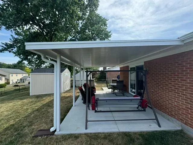 A white patio cover extends from a brick house, covering concrete patio with outdoor furniture.