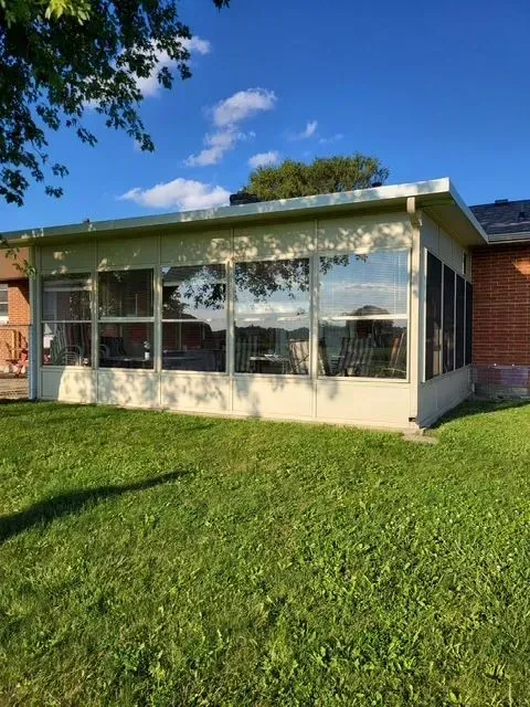 Tan-colored sunroom with large windows and screens, attached to a brick house, set on green grass under a blue sky.