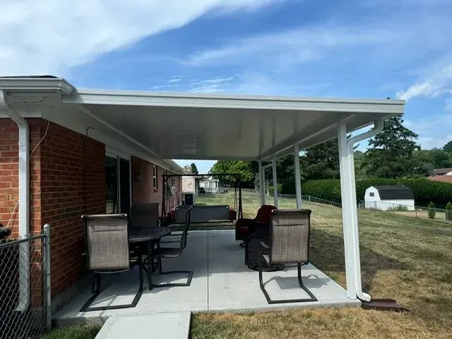 Patio with a white roof, chairs, and table. Brick house with a chain-link fence on a sunny day.