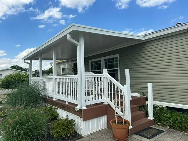 Covered porch with white railings and steps attached to a light green house. Blue sky overhead.