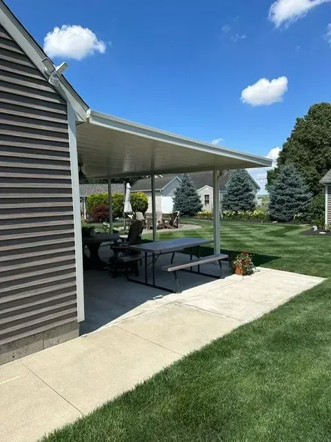 Patio with white roof attached to a house; table and chairs are underneath. Green lawn, blue sky.