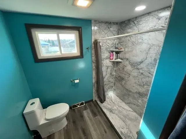 Bathroom with teal walls, gray shower, window, toilet, and gray wood-look flooring.