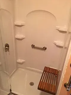 White shower stall with built-in shelves, a grab bar, and a wooden bench.