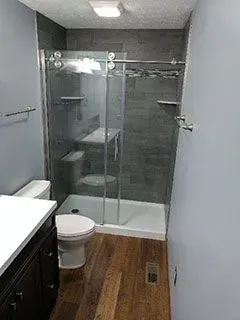 Bathroom with gray tile shower, sliding glass door, white toilet, vanity, and wood-look flooring.