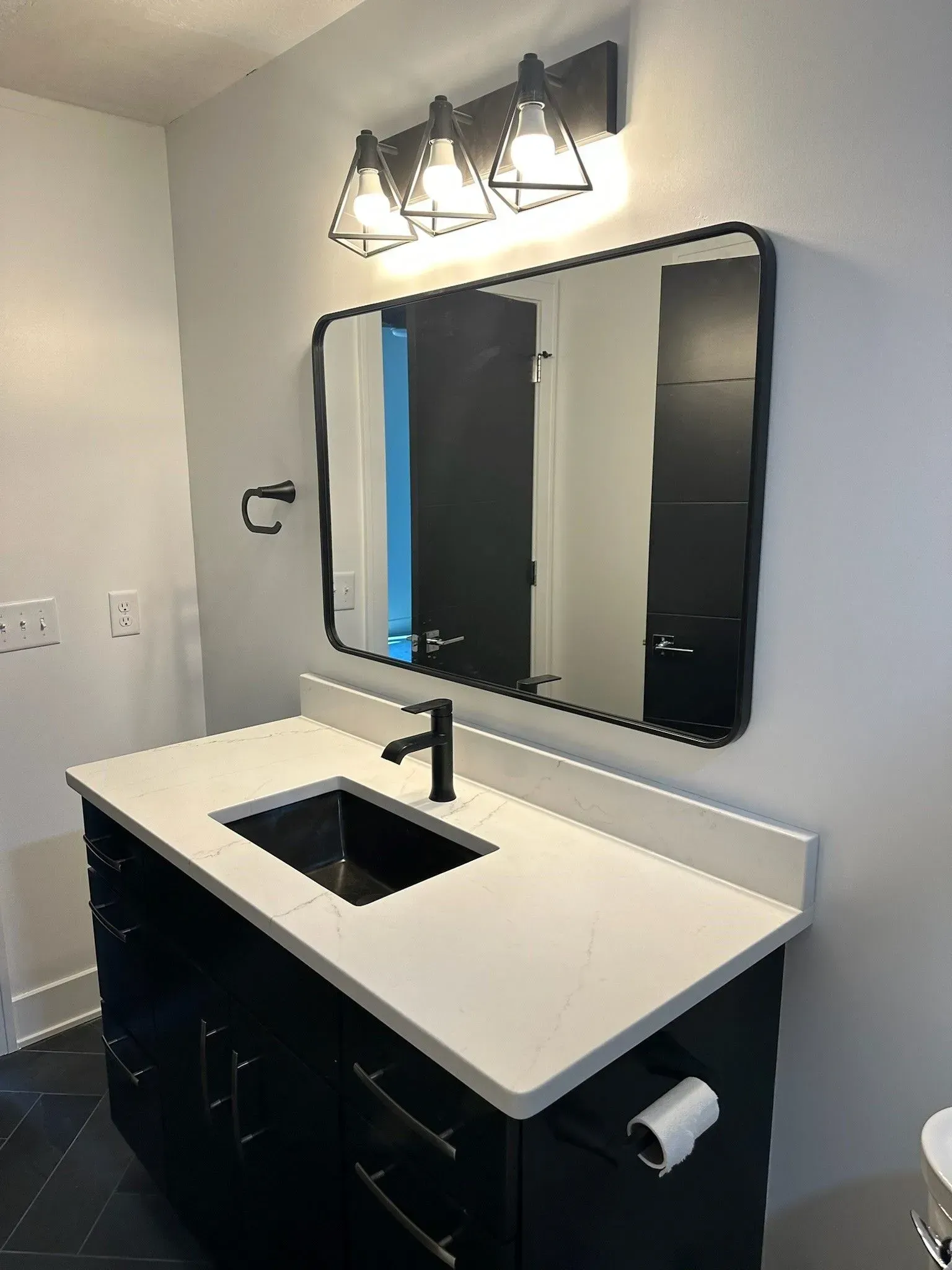 Bathroom with black vanity, white countertop, black sink, and mirror. White walls, black light fixture.