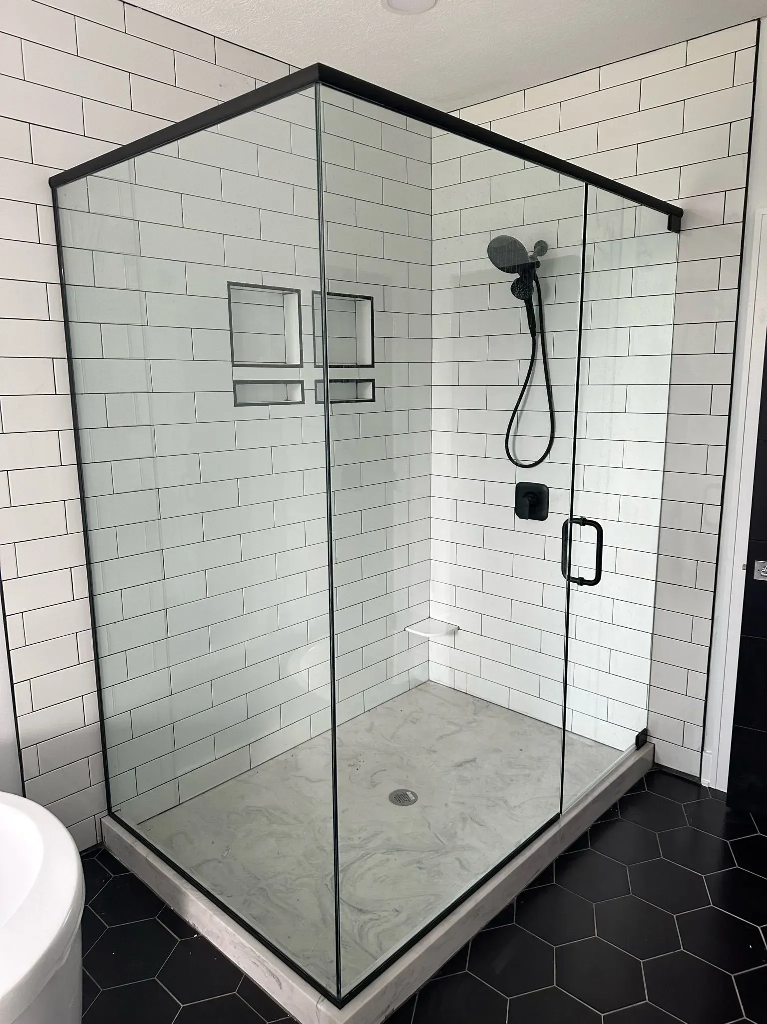 A corner shower with black trim and a glass door, inside a white-tiled bathroom with black hexagonal floor tiles.