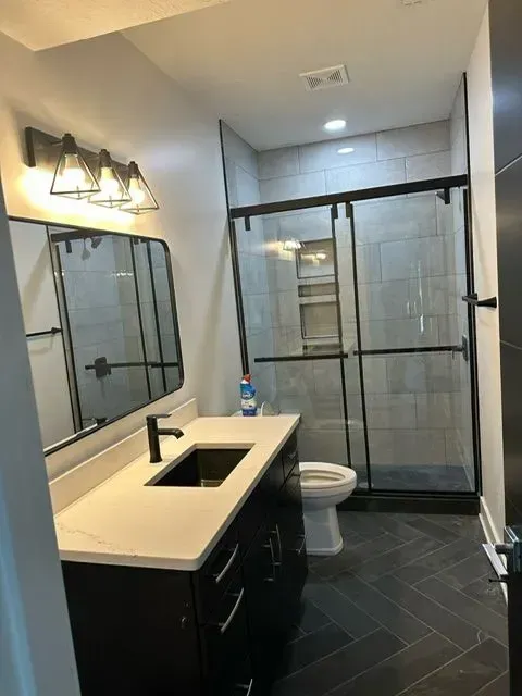 Modern bathroom with dark vanity, white countertop, glass shower, and gray tile.