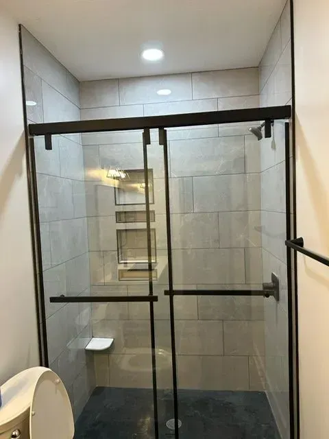 Modern bathroom shower with sliding glass doors, gray tiled walls, and dark metal hardware.