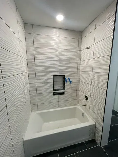 Bathroom with white textured tiles, built-in tub, recessed shelf, and black floor tiles.