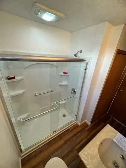 Bathroom with white shower stall, glass doors, and brown flooring. Sink and cabinet visible.