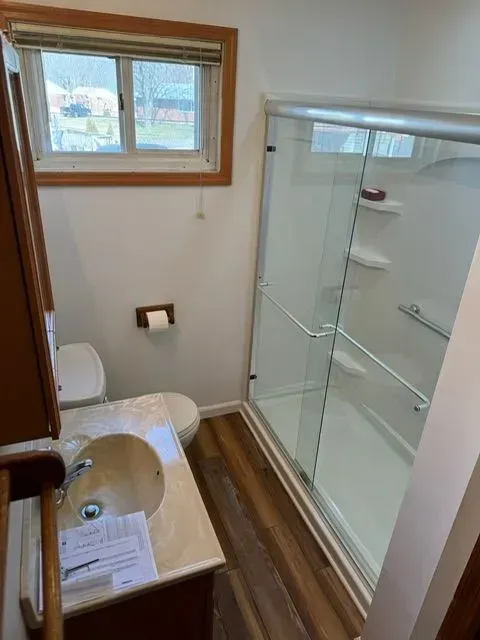 Bathroom with shower, toilet, and vanity. White walls, wood-look flooring, and a window above the toilet.