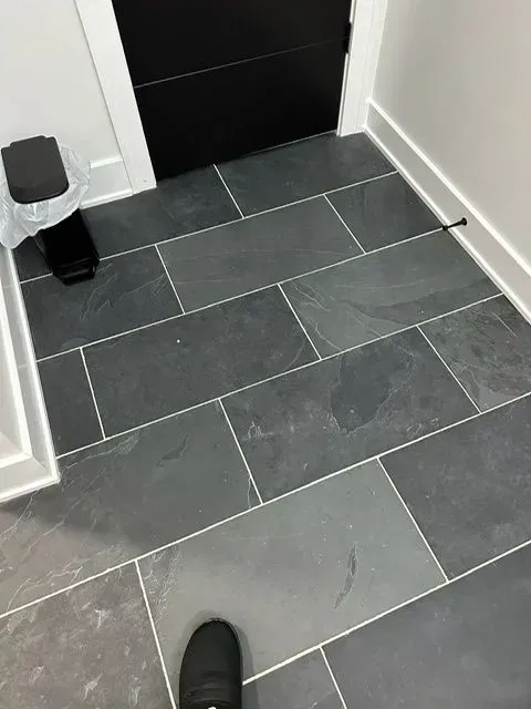 Dark gray rectangular floor tiles with white grout in a room. A black door, trash can, and foot are visible.