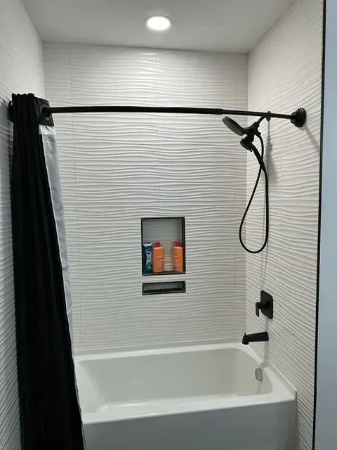 White tiled shower with black shower curtain, fixtures, and shampoo niche.