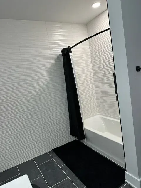 Bathroom with white textured tile walls, black floor, black shower curtain, and a white tub.