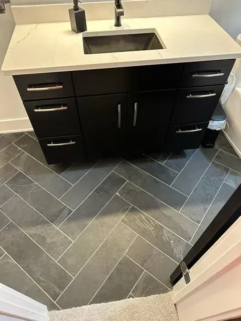 Black bathroom vanity with a white countertop and dark gray herringbone tile floor.