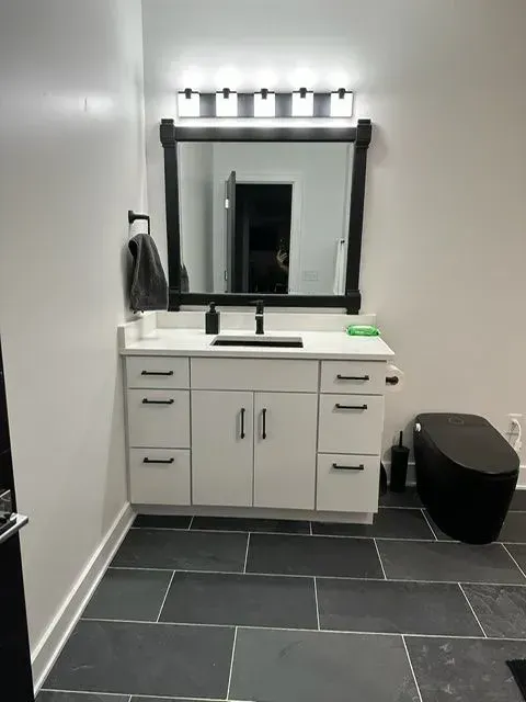 White bathroom vanity with black hardware, mirror, and toilet. Dark gray tiled floor.