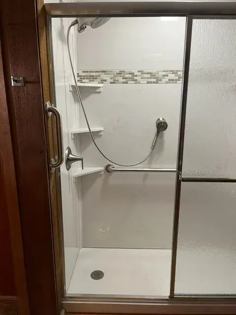 Accessible shower stall with sliding glass door, grab bar, showerhead, and built-in shelves.