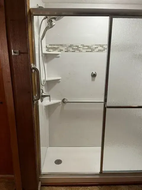 White shower stall with sliding glass doors, grab bar, built-in shelves, and a hand shower.