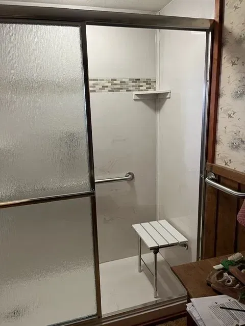 Accessible shower with sliding glass door, grab bar, shower seat, and small shelf.