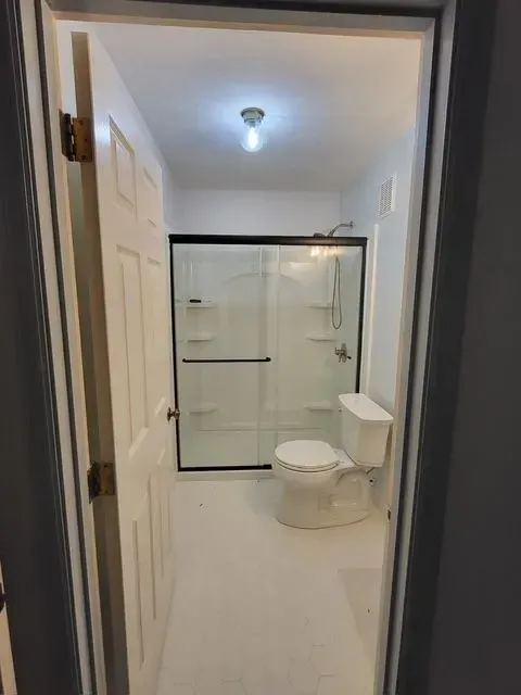 Bathroom interior, white walls and floor, black-framed shower, toilet, and partially open door.