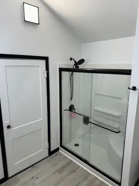 A white bathroom with a glass shower and black trim. A white door is on the left.