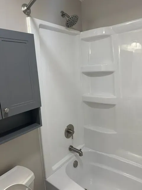 A white bathtub and shower enclosure with built-in shelves. Gray cabinet and fixtures are present.