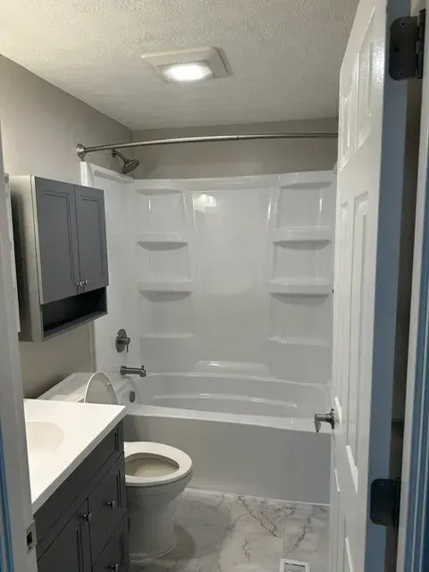 Bathroom with white tub, toilet, and vanity. Gray cabinet, white walls, and marble-look floor.