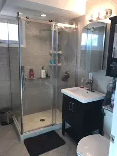 Bathroom with a glass shower, dark vanity, and toilet. Gray tile walls and black rug.