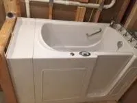 Walk-in bathtub installed against a wooden wall, with a silver grab bar and faucet.