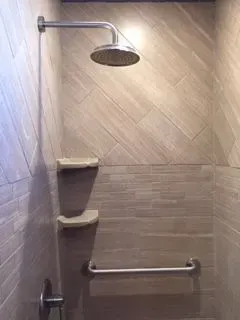 Shower with angled tile, overhead showerhead, two shelves, and a horizontal grab bar.