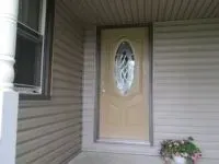 Beige front door with oval glass panel on a house with beige siding; a window and potted flowers visible.