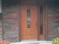 Brown wooden front door with stained-glass window and sidelight, set in a brick wall.