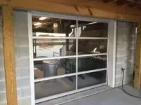 Glass garage door in a concrete block wall. Reflects items in a workshop, including a trash can and a metal table.