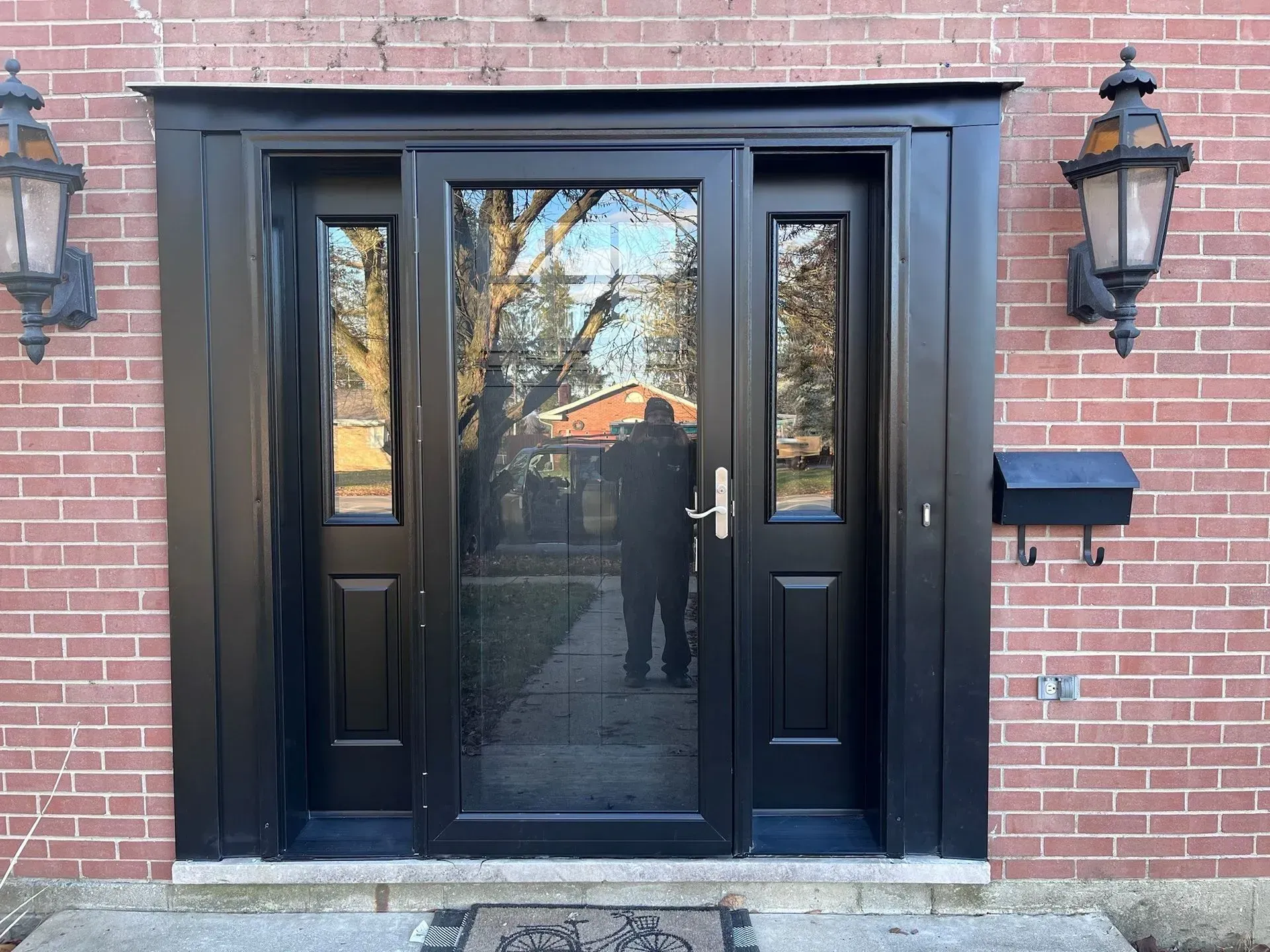 Black front door with sidelights, set in brick facade, flanked by sconces and mailbox.
