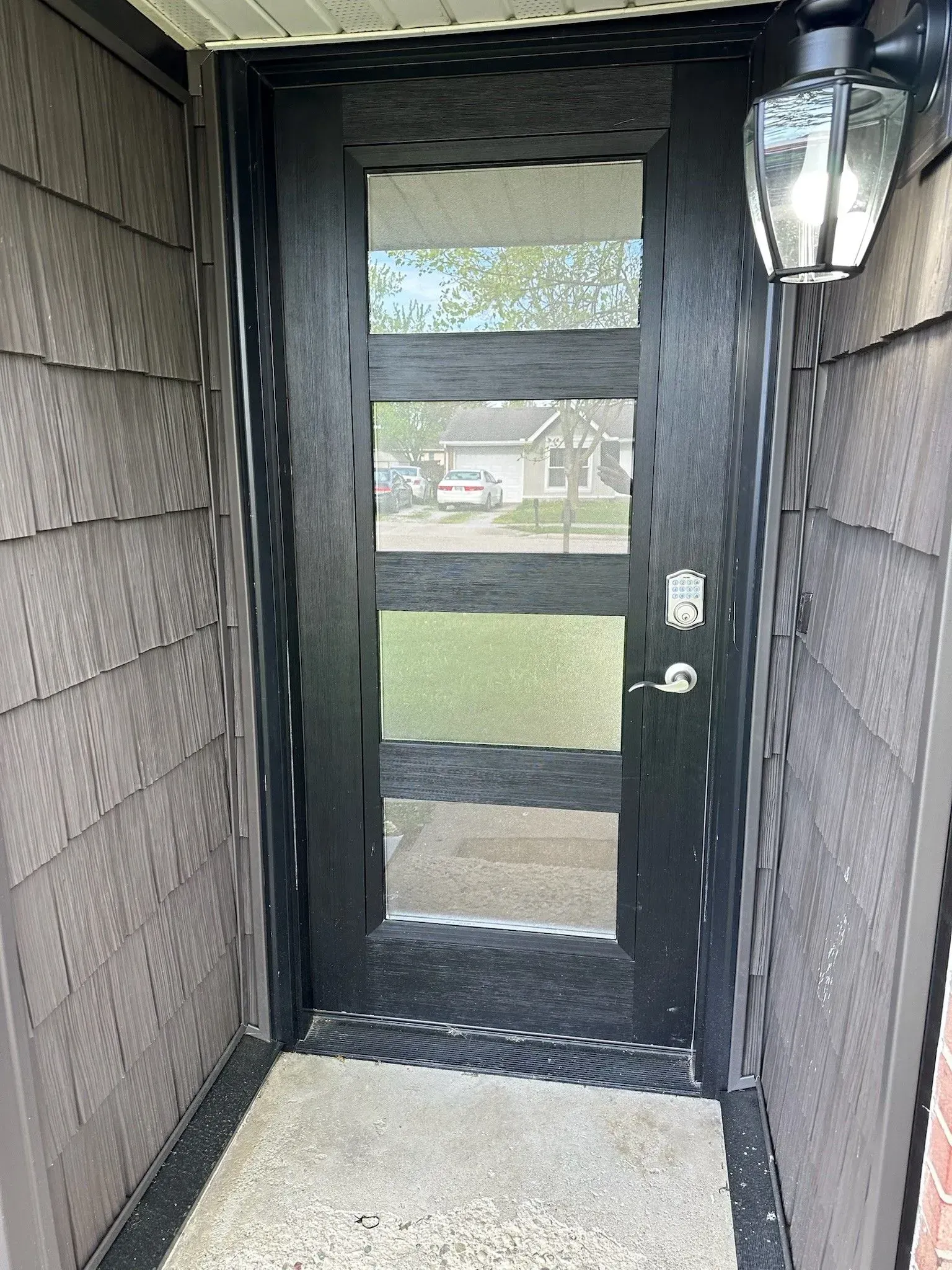 Black front door with horizontal glass panes, gray siding, and porch light.