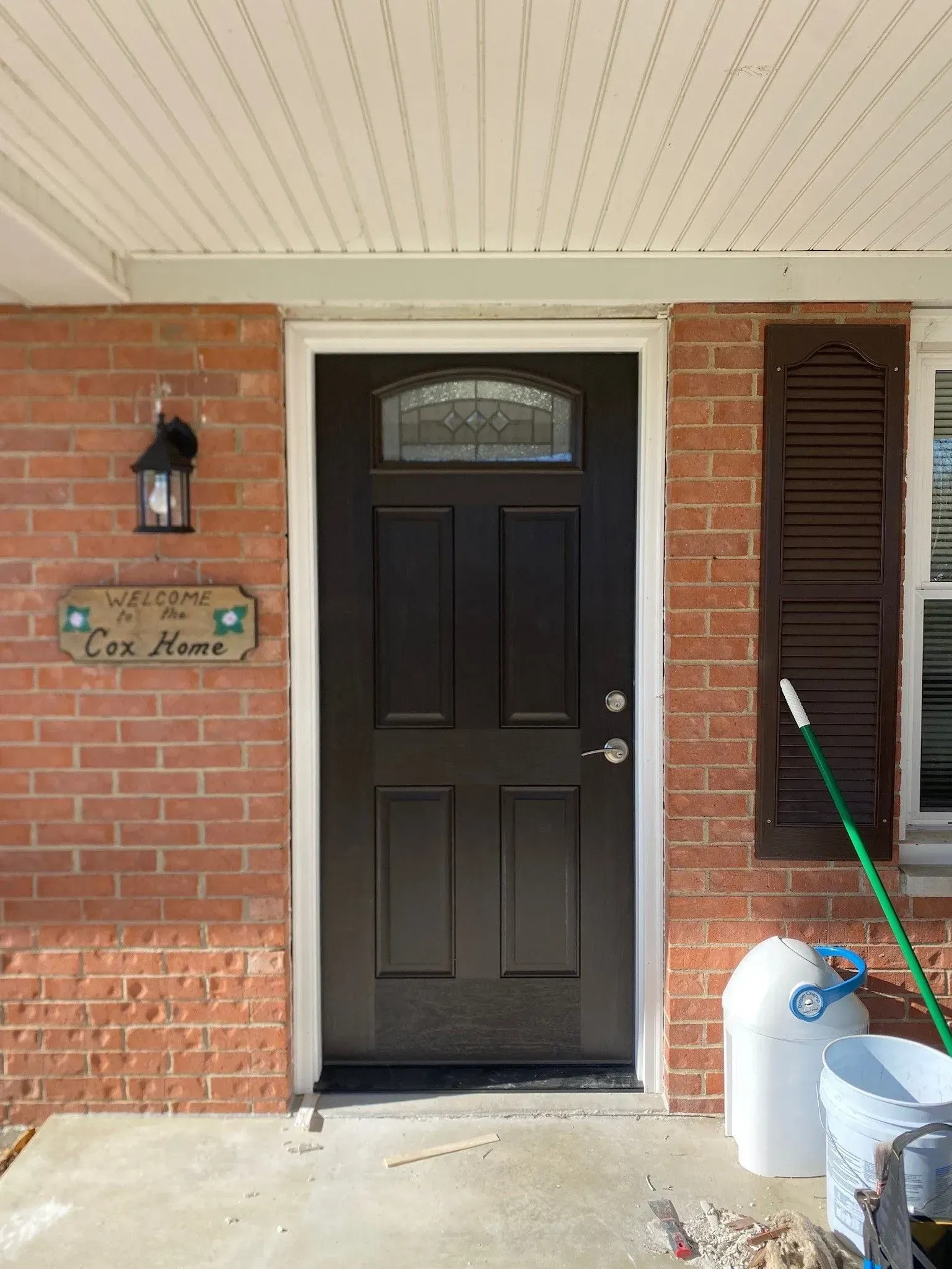 Brick entryway with dark brown door, sidelight, and shutters; welcome sign; outdoor light fixture.