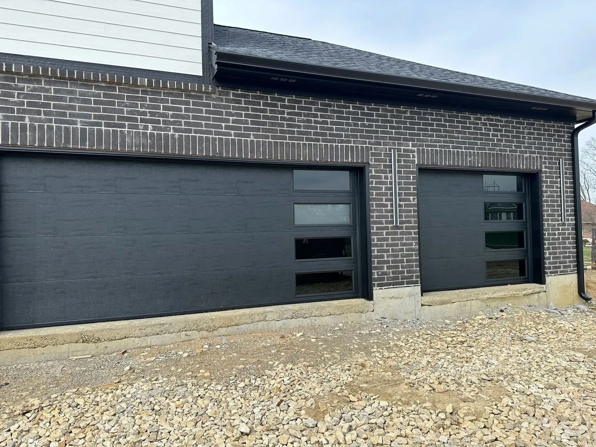 Two black garage doors with glass panels on a brick building with gravel driveway.